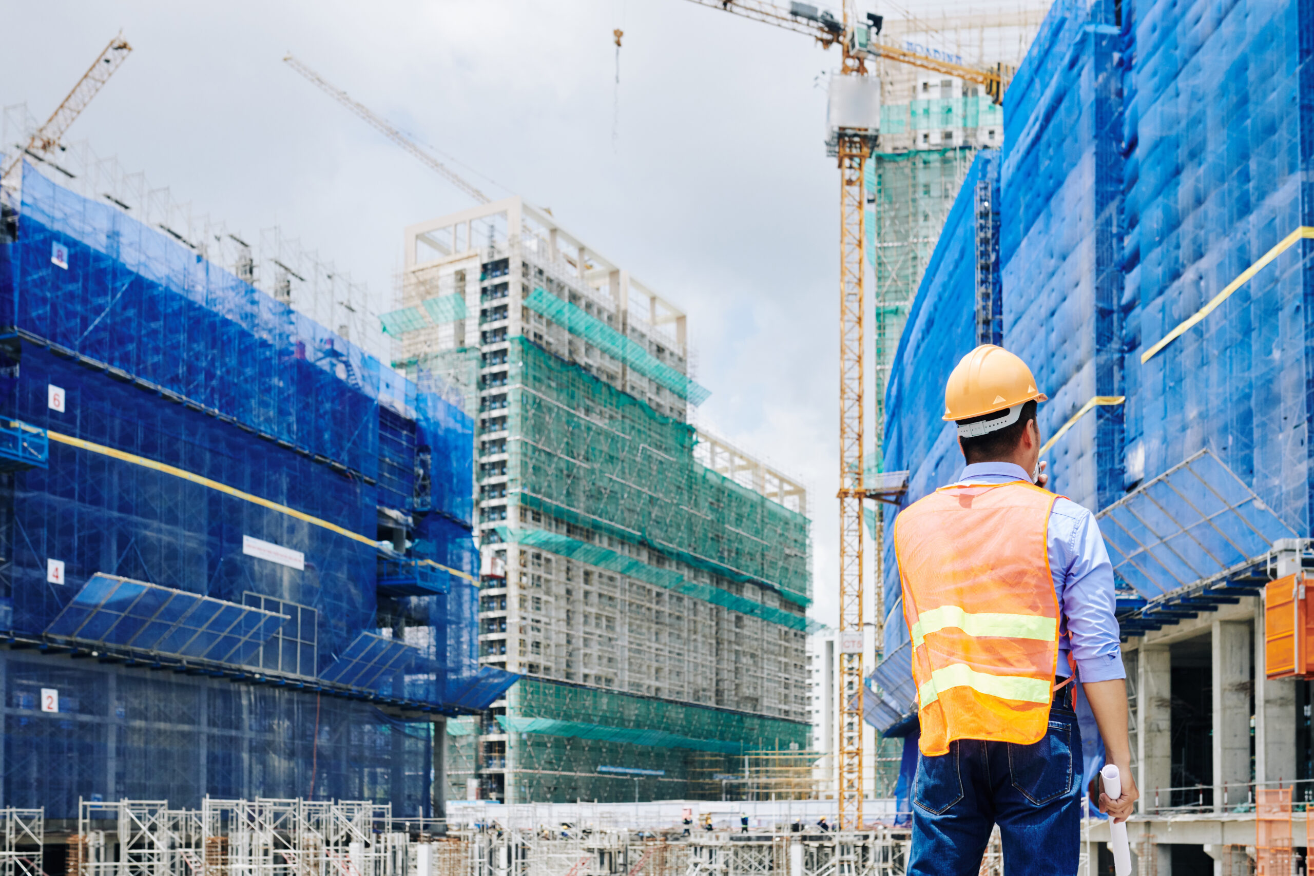 Rear view of professional engineer in bright orange vest talking on walkie-talkie when looking at building under construction